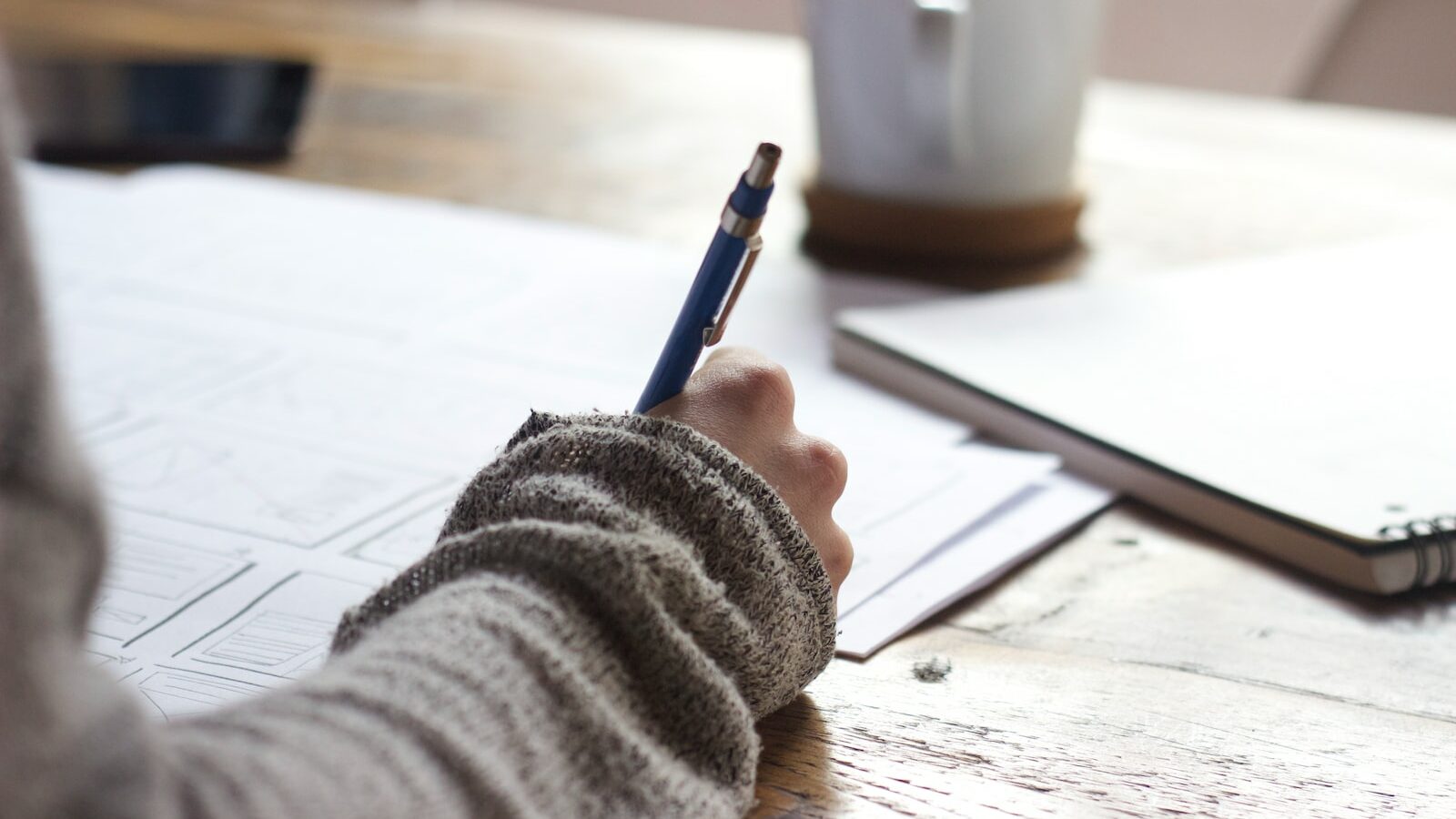 person writing on brown wooden table near white ceramic mug